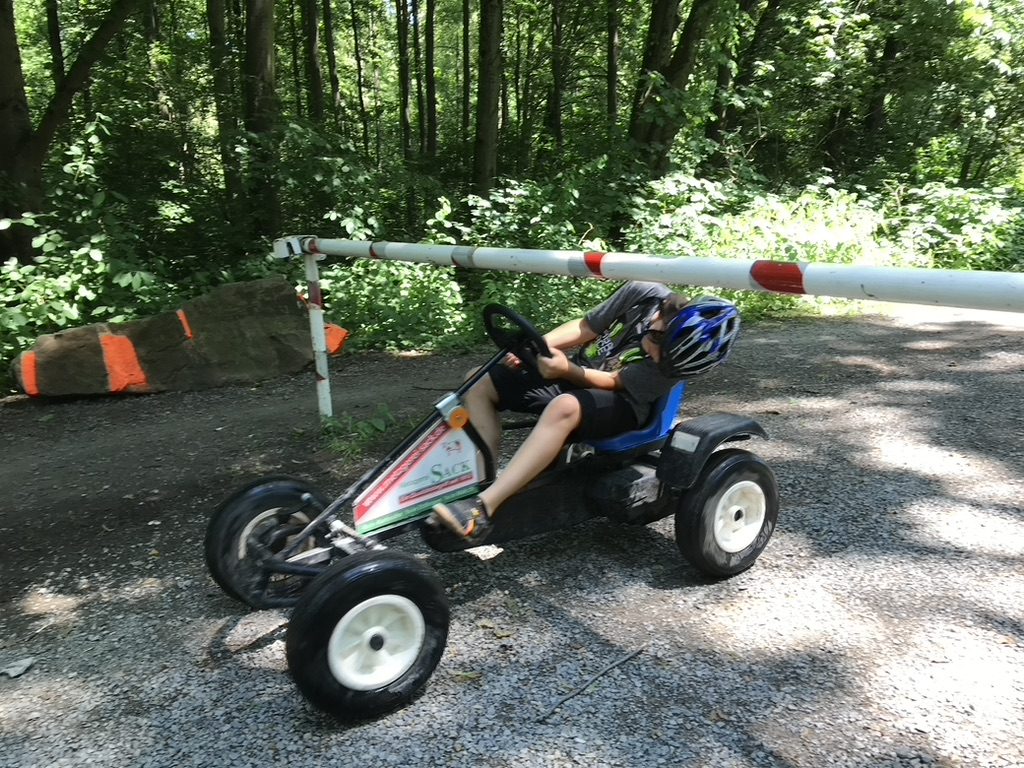 Junge fährt mit einem Pedal-Kett-Kart mit Werbeschild an einer Schranke im Wald vorbei